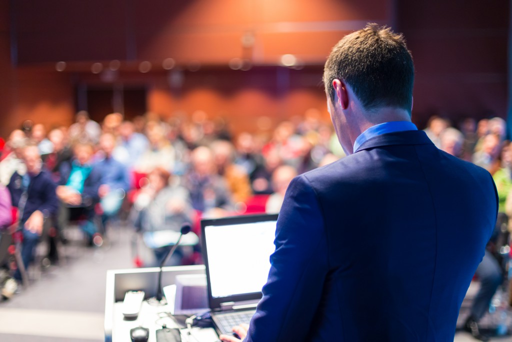 A man in a suit looks at a laptop atop a lectern as he speaks to an audience.